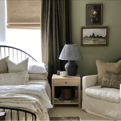 A bedroom with a bed with a white comforter next to a nightstand and white chair.