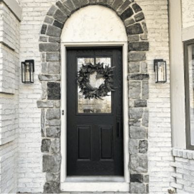 An outdoor entryway to a white brick house with a black painted front door with a natural wreath hanging in front of the window. Credit: Instagram @konapropainting