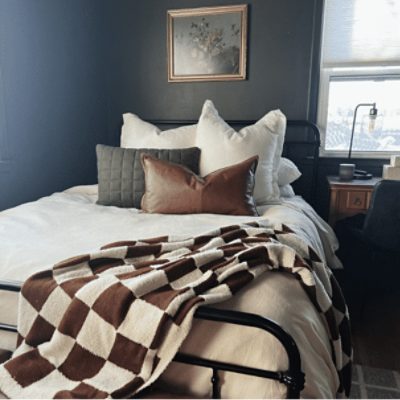 A dark blue painted bedroom with a wrought iron bed with neutral bedding next to a window. Credit: Instagram @mostlywanderinghome