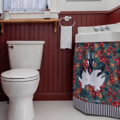 A bathroom with a white toilet and a wood shelf above next to a white sink with a curtain with swans on it. The wainscoting is done in SW 2802 Rookwood Red. ne