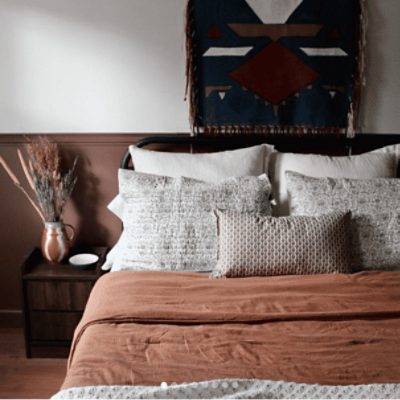 A bedroom with a bed with pillows and an orangish tan comforter. A wood nightstand with a vase and plant. An art piece hangs behind the bed on a white wall.