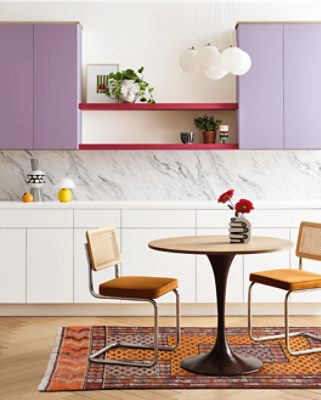 A modern kitchen with upper cabinets in Radiant Lilac, white marble backsplash, and white lower cabinets. A round table with red flowers sits on a colorful rug, with pendant lights above.