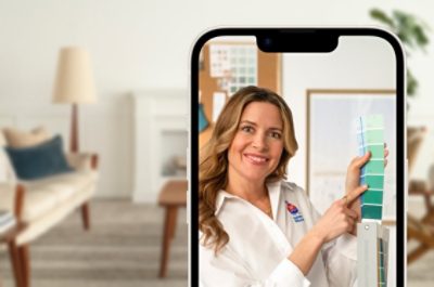 A phone showing a virtual color consultation with a woman holding up a fan deck. A white living room is blurred out behind the phone.