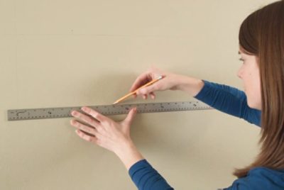 Person using a metal ruler to draw a straight horizontal line on a wall with a pencil for a precise measurement task.