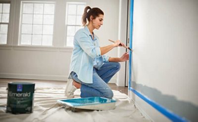 A woman painting around a door using a paint brush and tray.