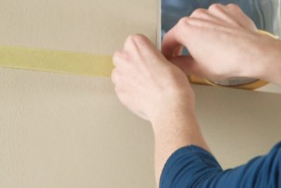 Close-up of hands applying yellow masking tape to secure the edge of a poster or paper on a beige wall.