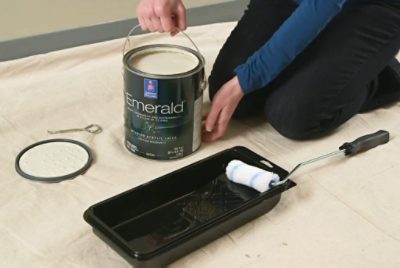 Person kneeling on a drop cloth with an open can of Emerald interior paint, preparing to paint with a roller and tray nearby.