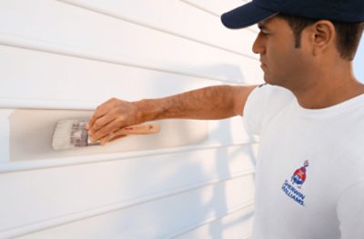 A person wearing a white shirt with the Sherwin-Williams logo painting siding on a house with a brush.