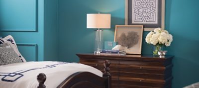 A pale blue bedroom wall with dresser, wall art, and a bed