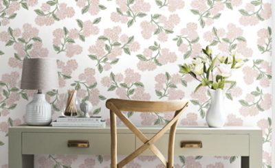 Styled home office vignette featuring floral peel-and-stick wallpaper with blush pink blossoms and sage green leaves, a light wood chair, gray table lamp, decorative books, and a white vase of flowers on a painted desk.