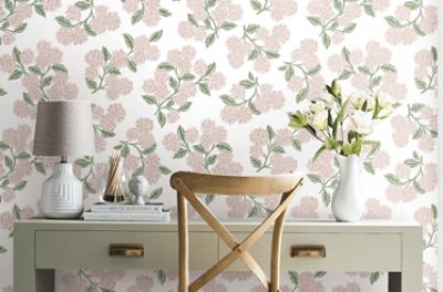 Styled home office vignette featuring floral peel-and-stick wallpaper with blush pink blossoms and sage green leaves, a light wood chair, gray table lamp, decorative books, and a white vase of flowers on a painted desk.
