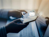 Person using a smartphone while working on a laptop, with floating icons above the laptop representing phone, envelope, and email communication.